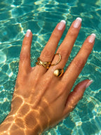 Bague croisée en acier inoxydable doré au design ouvert et aux lignes entrelacées porté sur une main qui touche l'eau