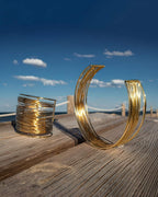 ensemble collier et manchette dorée en acier inoxydable large en forme de spirales posés sur une table en bois en bord de mer