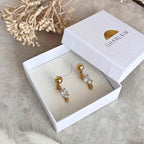 Boucles avec une boule au niveau du lobe et un anneau fin qui revient de derrière jusqu’à l’avant, serti d’un diamant présentées dans un joli écrin blanc au nom de la marque